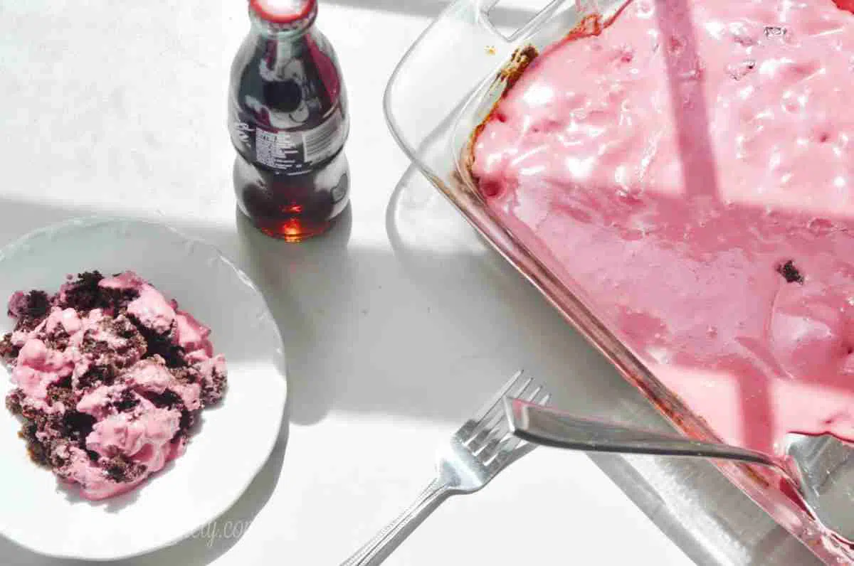 cherry coke earthquake cake on a white plate, with a glass bottle of coca cola and a baking dish of cake.
