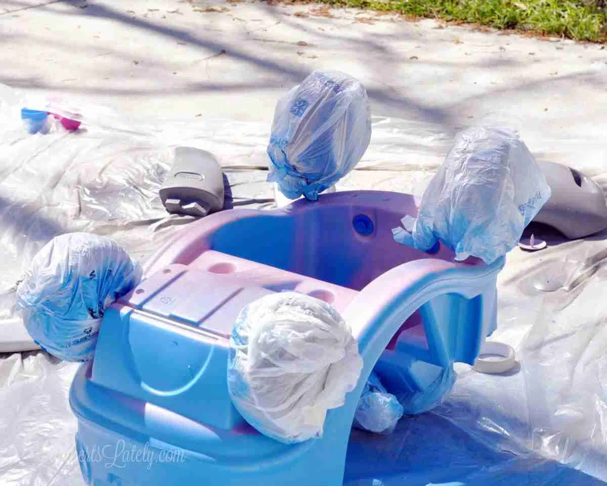 cozy coupe base turned upside down, wheels wrapped in plastic shopping bags, while painting blue.