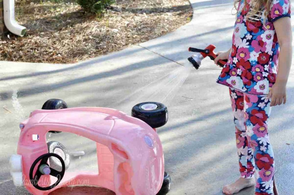 little girl spraying cozy coupe with hose.