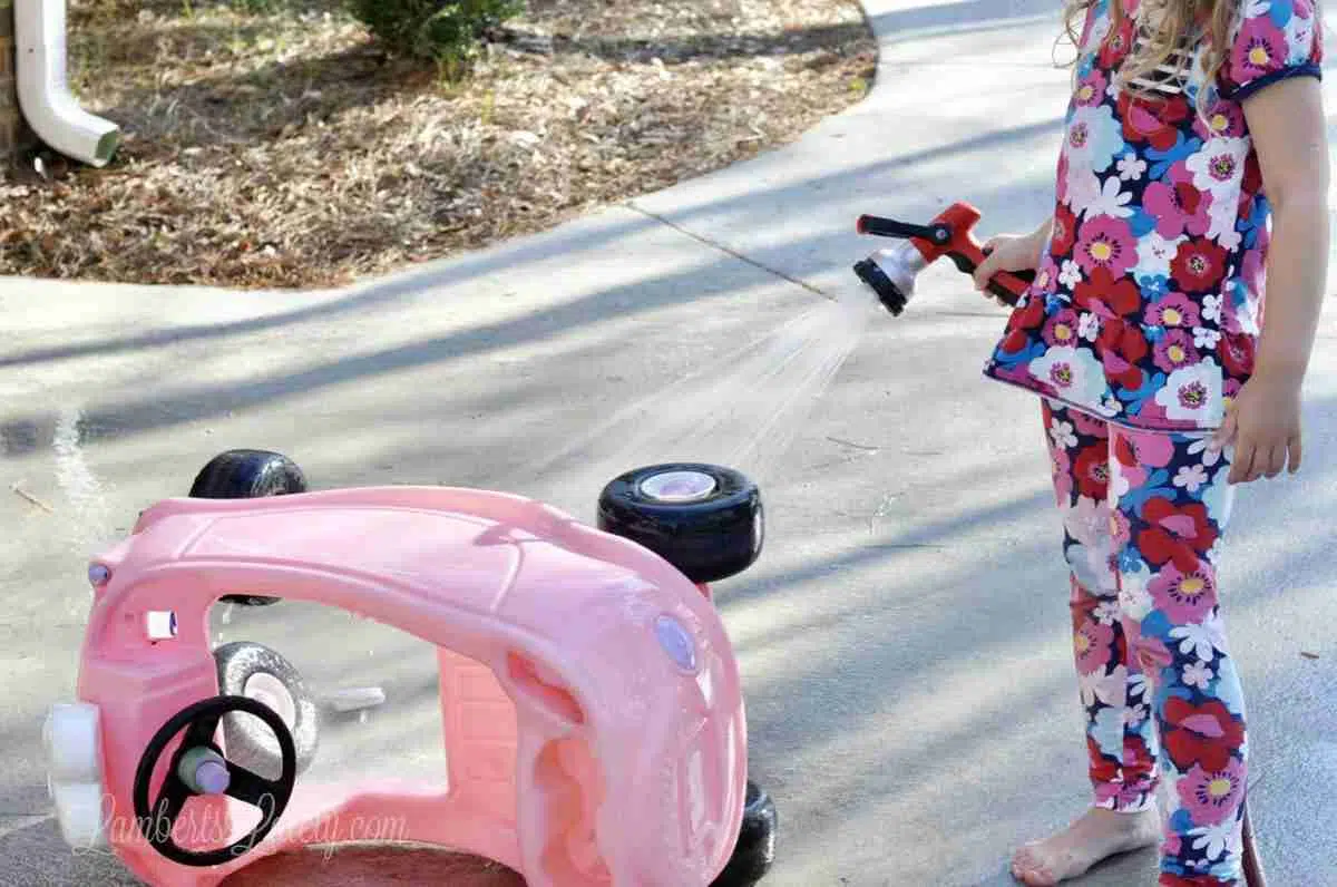 little girl spraying cozy coupe with hose.