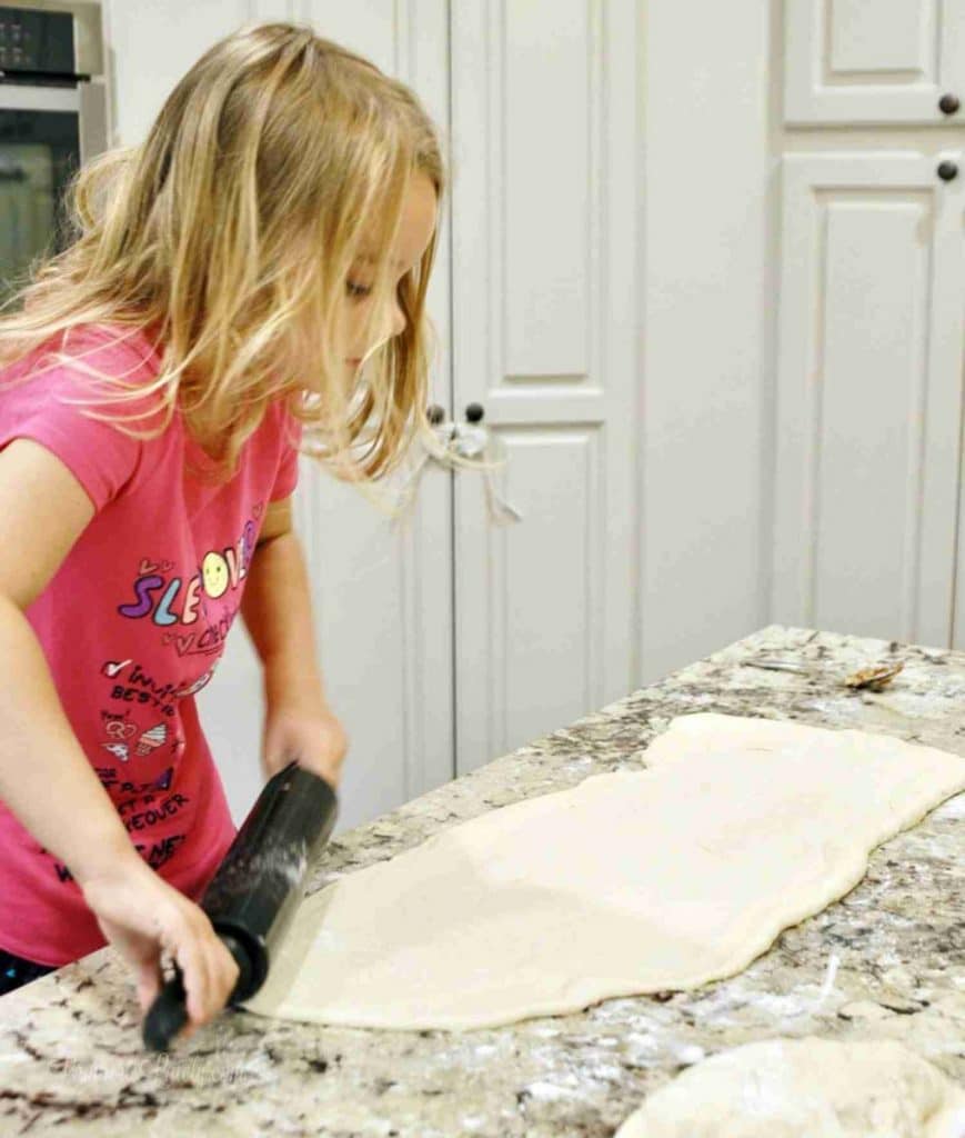 Child rolling out dough for cinnamon rolls in a kitchen with white cabinets.