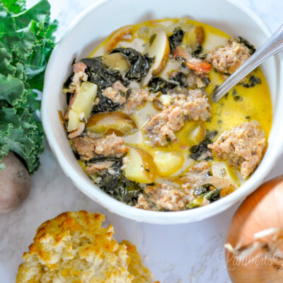 bowl of zuppa toscana next to bunch of kale, onion, garlic, and cheddar biscuit.