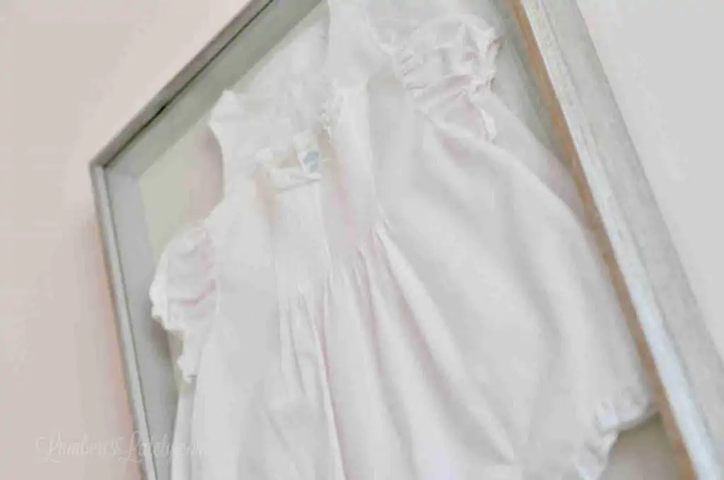looking up on a pink baby dress hanging on a pink wall in a shadow box.