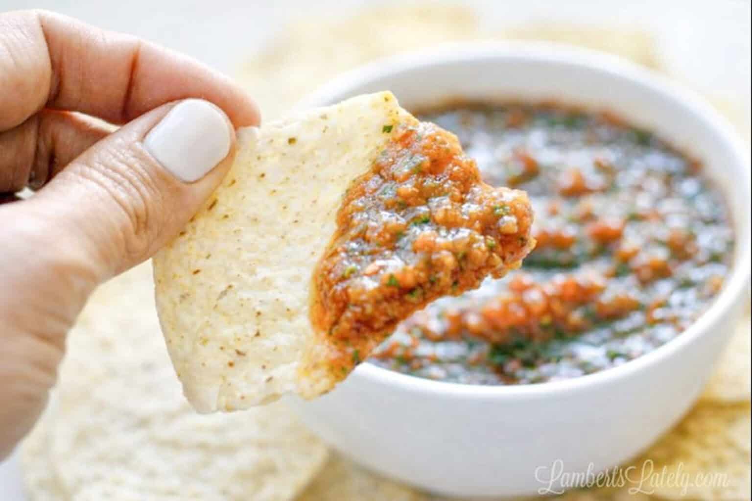 Close-up of a hand dipping a tortilla chip into salsa in a white bowl.