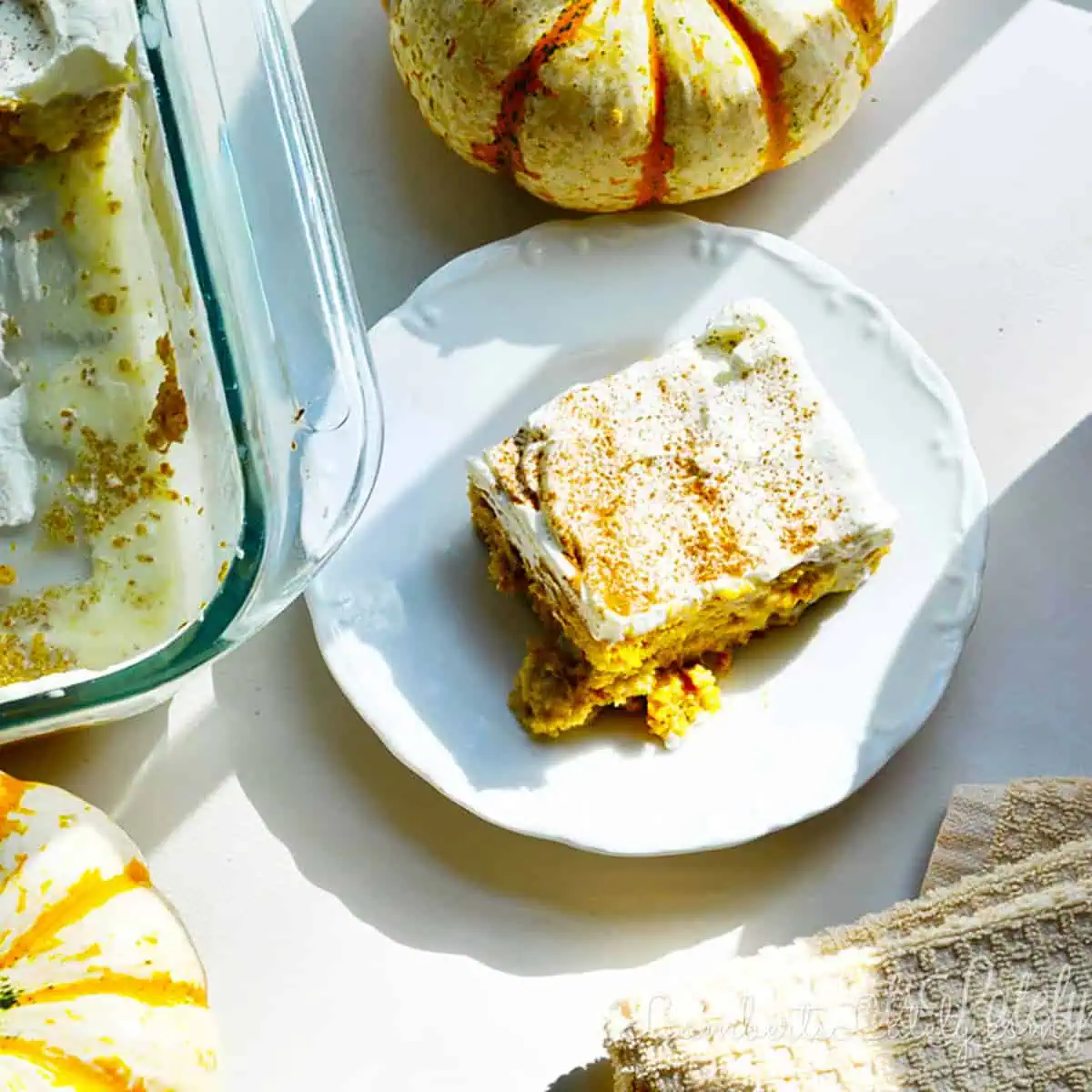 piece of pumpkin tres leches cake on a plate next to a baking dish and pumpkins.