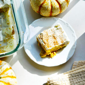 piece of pumpkin tres leches cake on a plate next to a baking dish and pumpkins.