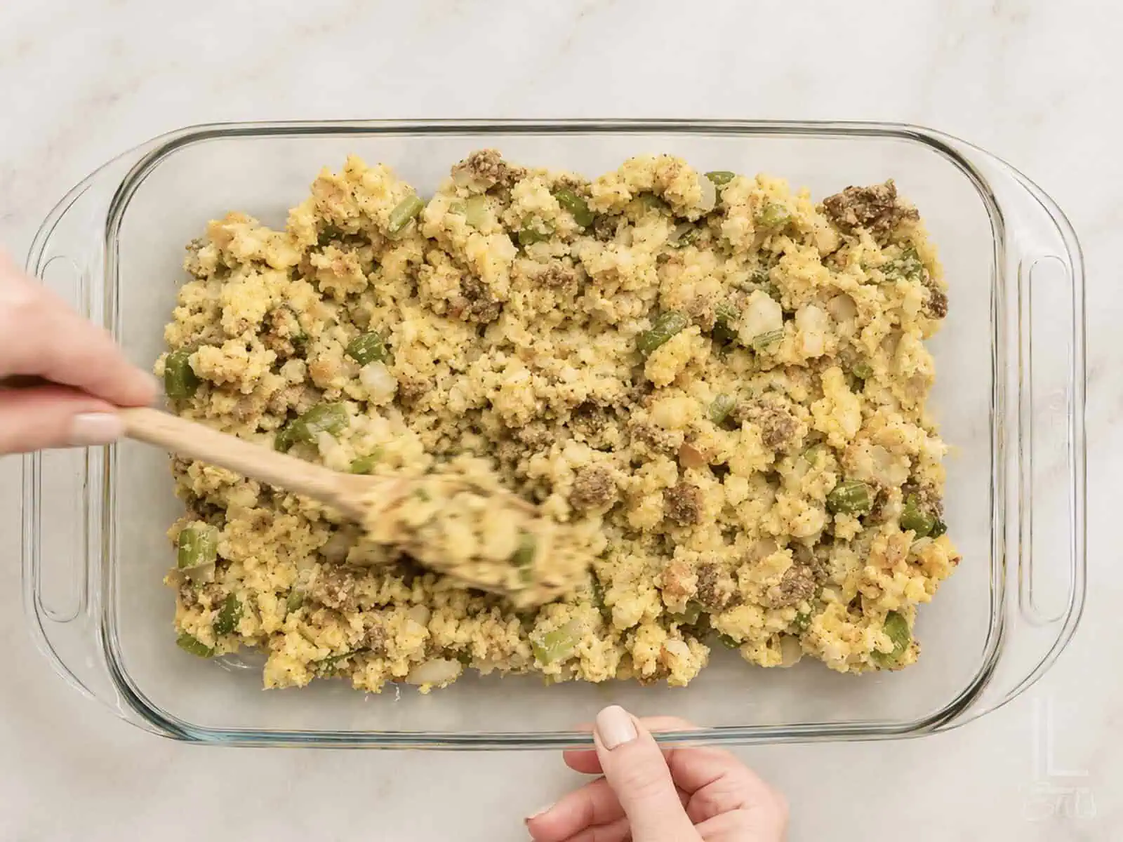 Unbaked Jiffy cornbread dressing with sausage in a glass dish, being spread for baking.