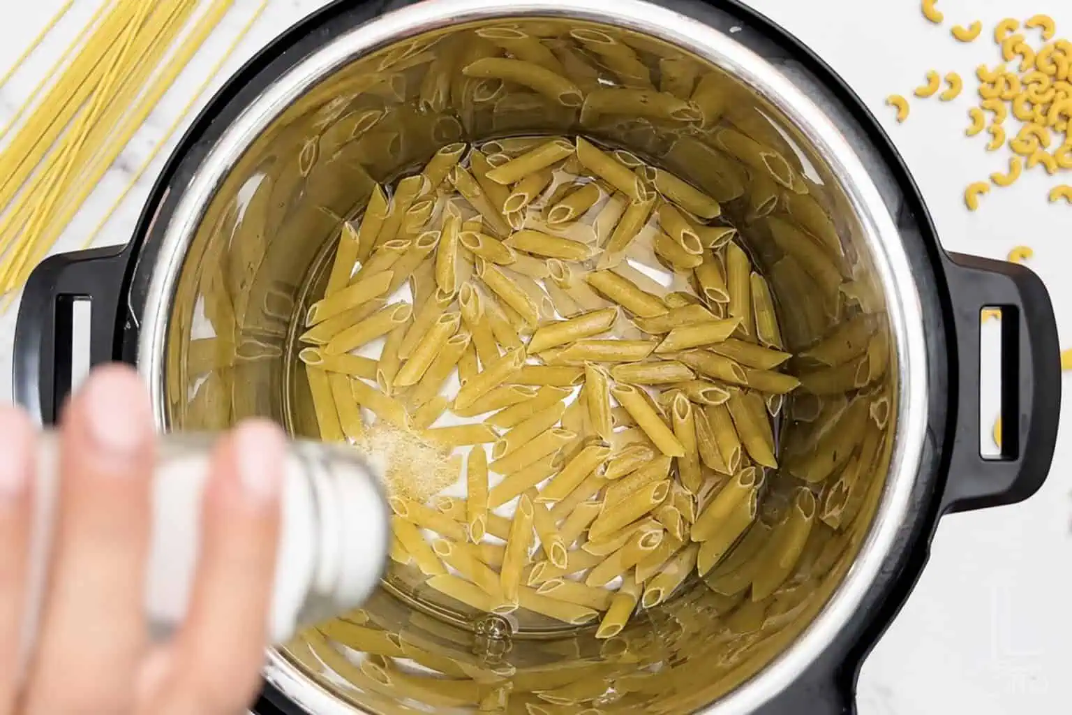 Uncooked pasta in an Instant Pot with a hand adding salt.