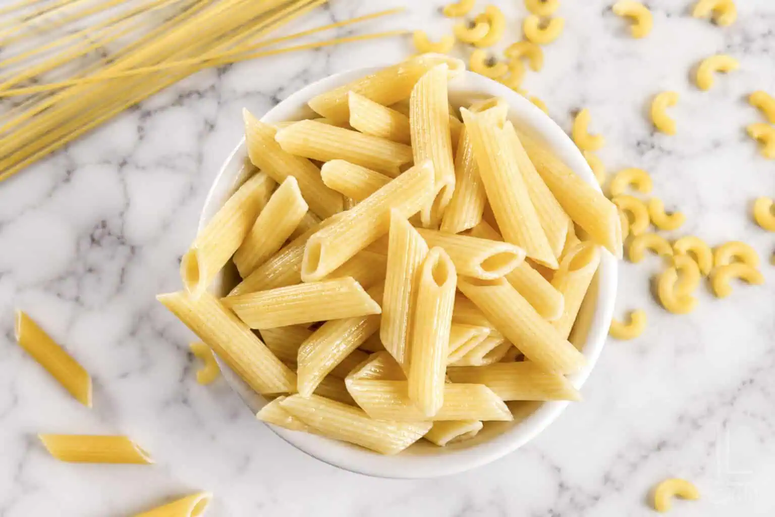 Cooked penne pasta in a white bowl on marble surface with uncooked pasta around.
