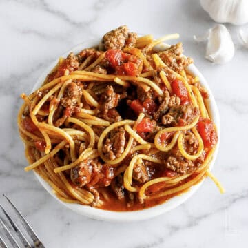 Bowl of spaghetti with meat sauce and tomatoes on a white marble surface.