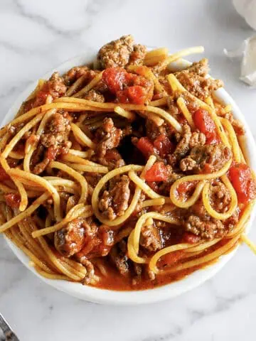 Bowl of spaghetti with meat sauce and tomatoes on a white marble surface.