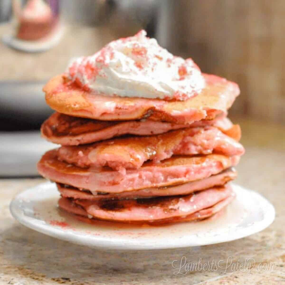 strawberry cake mix pancakes stacked on a plate, covered with icing syrup, whipped cream, and sprinkles.