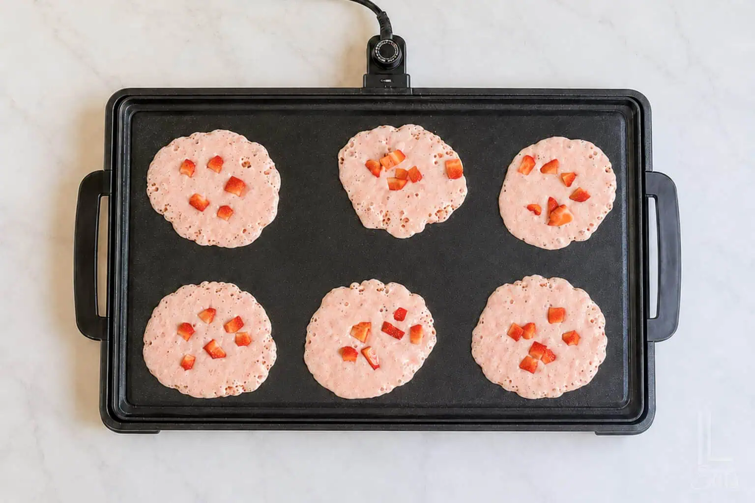 Six pink pancake batter portions with red strawberry pieces on a griddle.