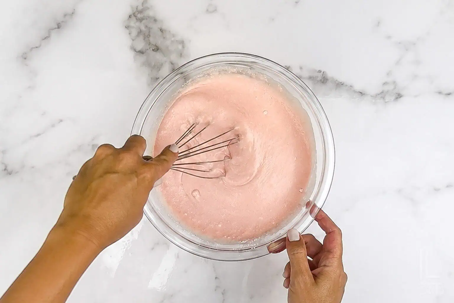 Mixing pink pancake batter in a glass bowl with a whisk on a marble surface.