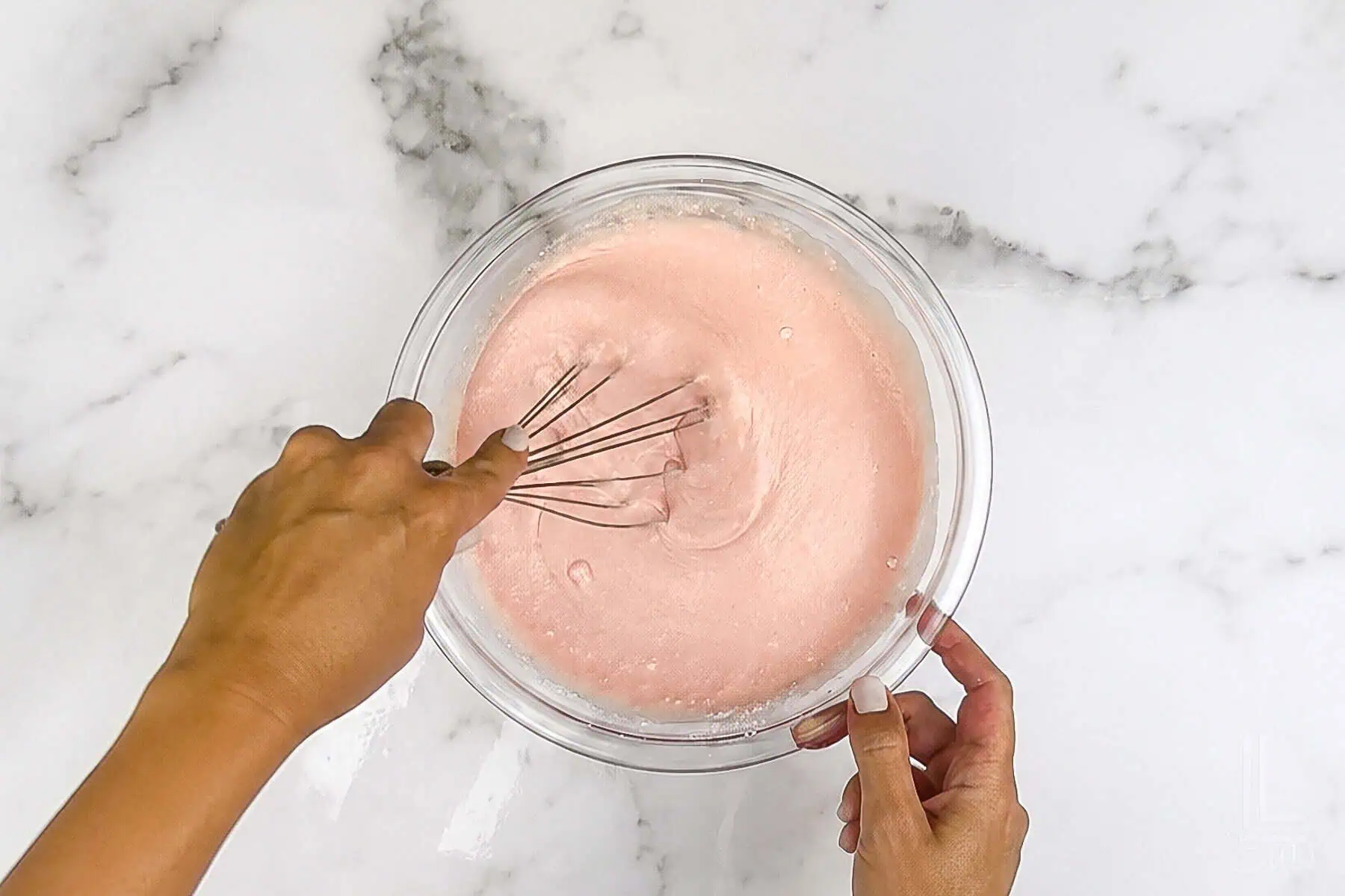 Mixing pink pancake batter in a glass bowl with a whisk on a marble surface.