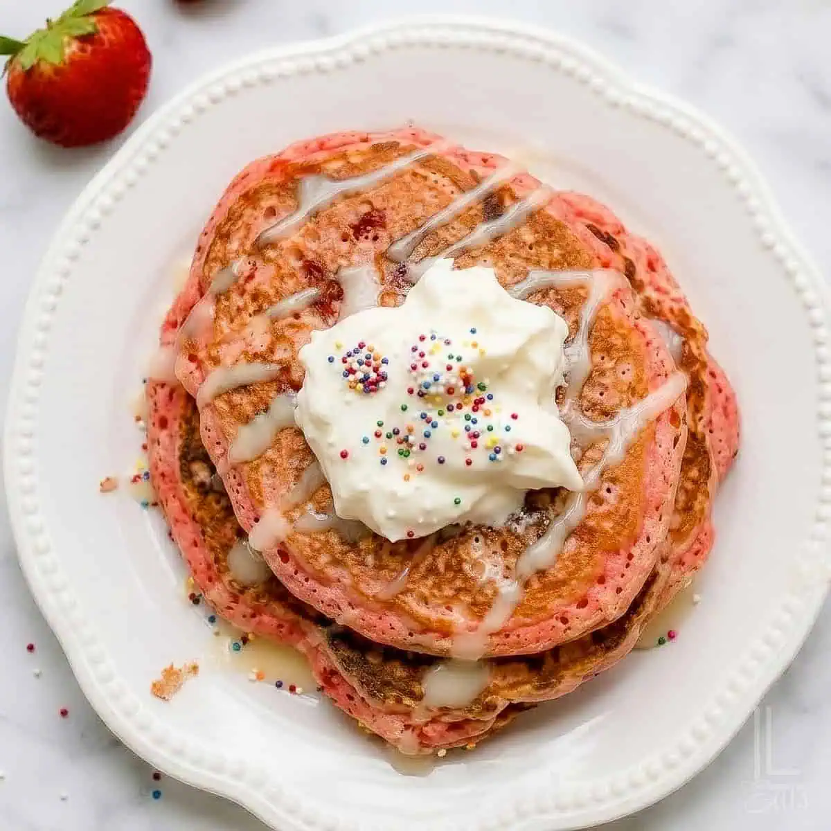 Stacked pink cake mix pancakes with icing syrup and sprinkles on a white plate.