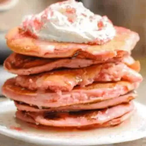 strawberry cake mix pancakes stacked on a plate, covered with icing syrup, whipped cream, and sprinkles.