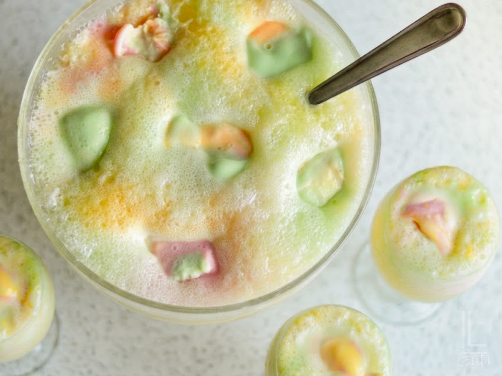 An overhead view of a festive punch bowl filled with rainbow sherbet punch alongside filled serving glasses.