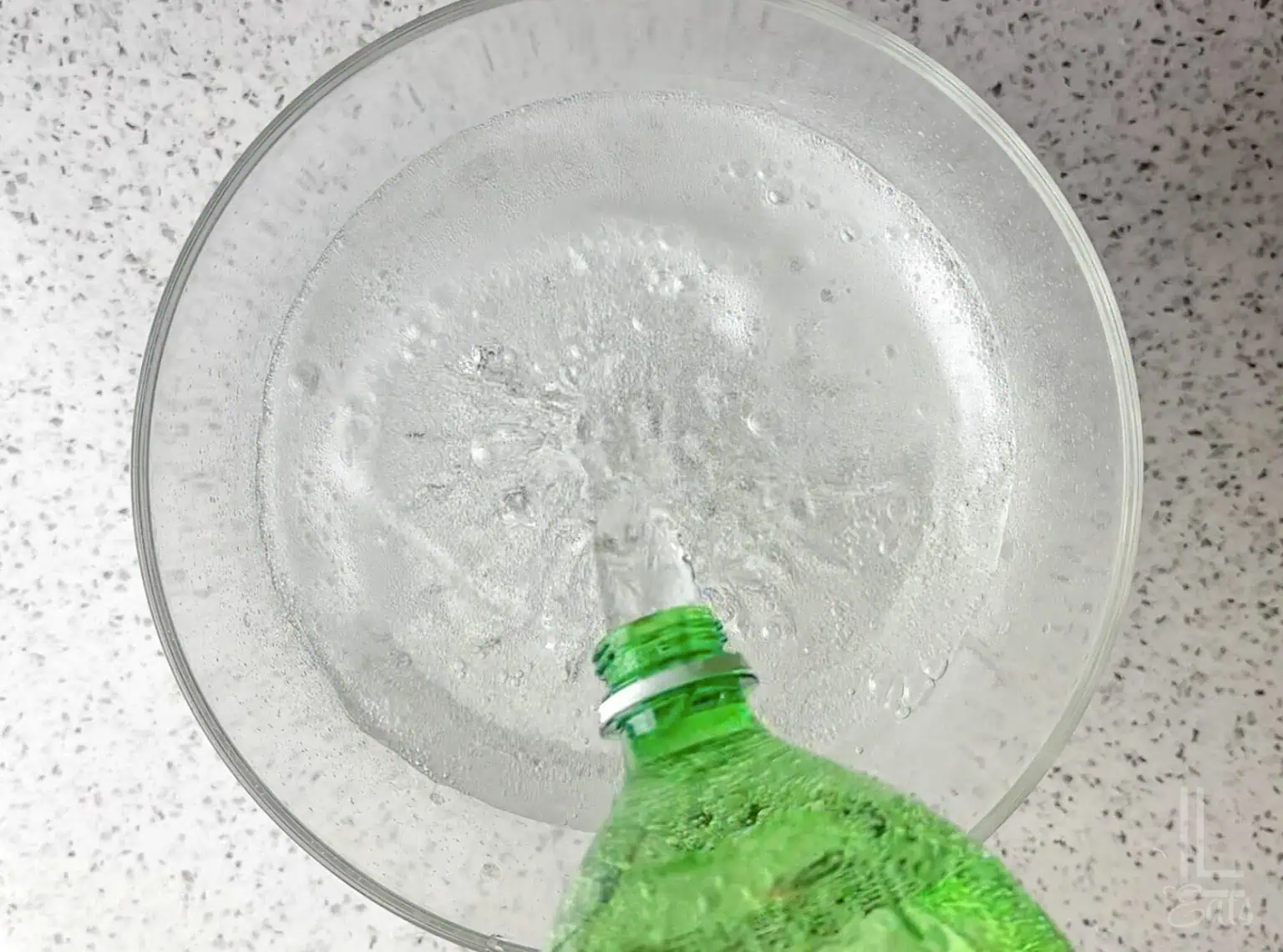Pouring a green bottle of lemon-lime soda (possibly Sprite) into a clear glass punch bowl.