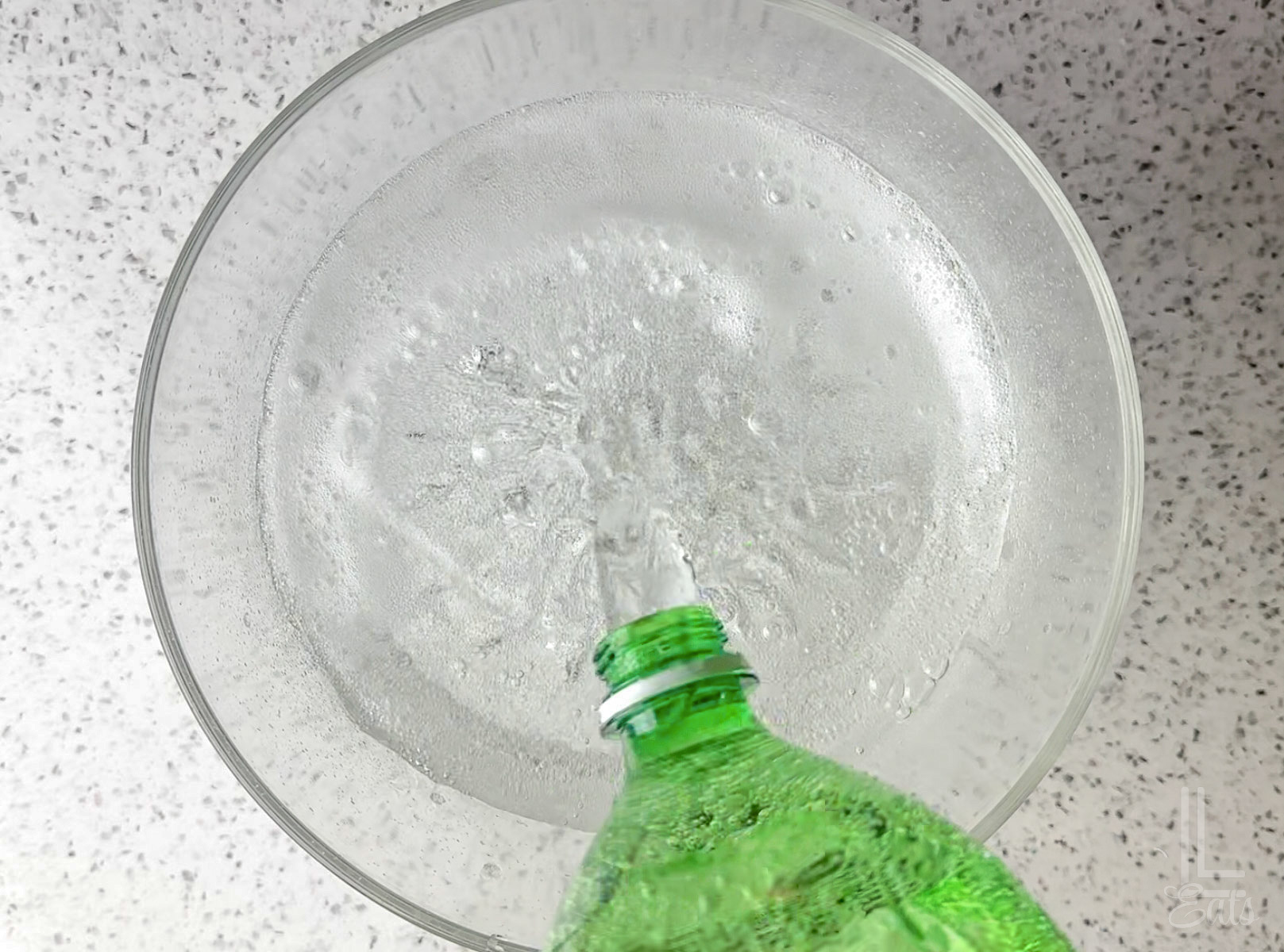 Pouring a green bottle of lemon-lime soda (possibly Sprite) into a clear glass punch bowl.