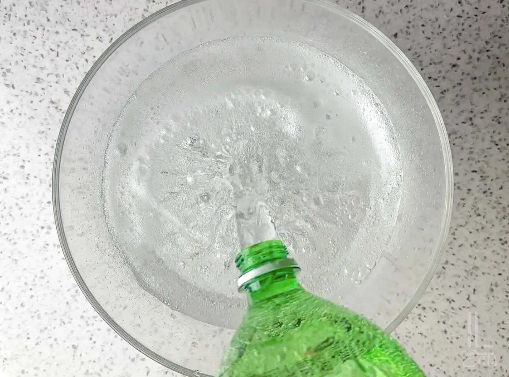 Pouring a green bottle of lemon-lime soda (possibly Sprite) into a clear glass punch bowl.
