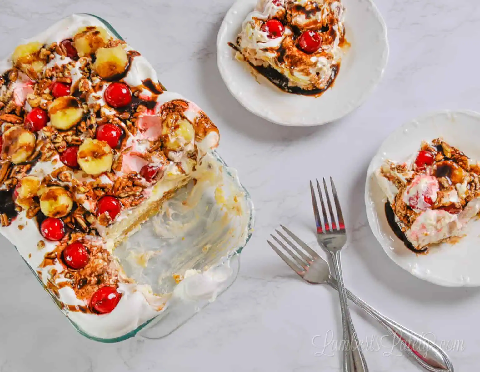 a layered banana split cake in a glass in a baking dish, topped with bananas, cherries, and chocolate syrup, with two plates and servings of the cake, and forks.
