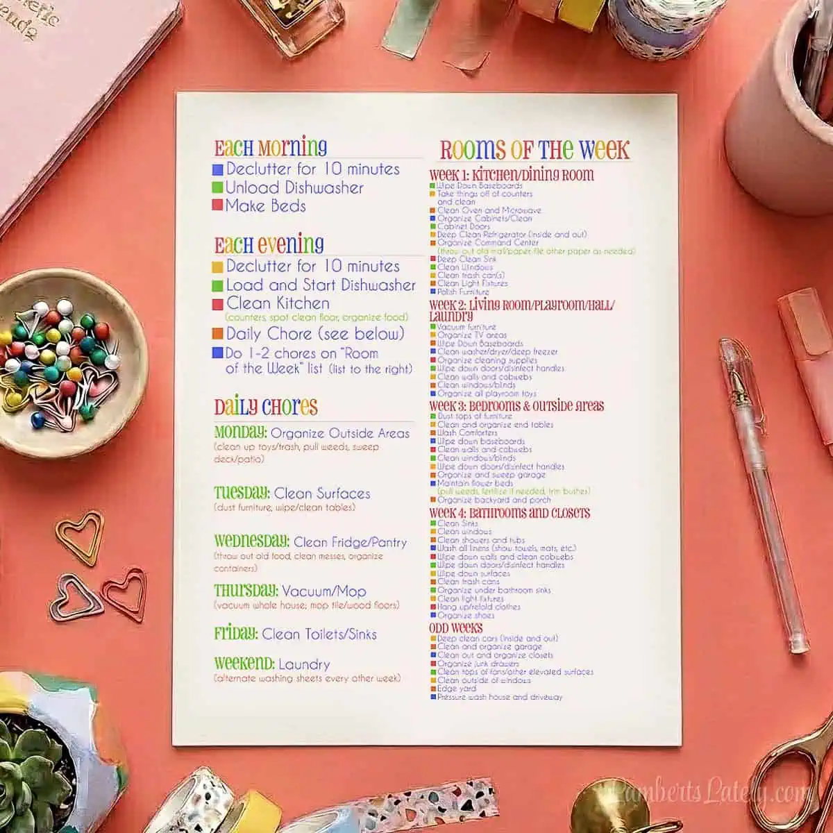 A top-down flat lay photo on a coral-pink background featuring a white cleaning checklist printable page surrounded by colorful office supplies, washi tape, scissors, markers, a stapler, and a small succulent plant.