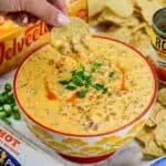 Close-up of a hand dipping a tortilla chip into creamy Rotel sausage dip in a bowl.