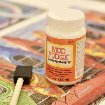 bottle of mod podge and a sponge brush on a finished jigsaw puzzle.