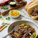 Delicious shredded Italian beef served on a plate with pickles and bread in the background.