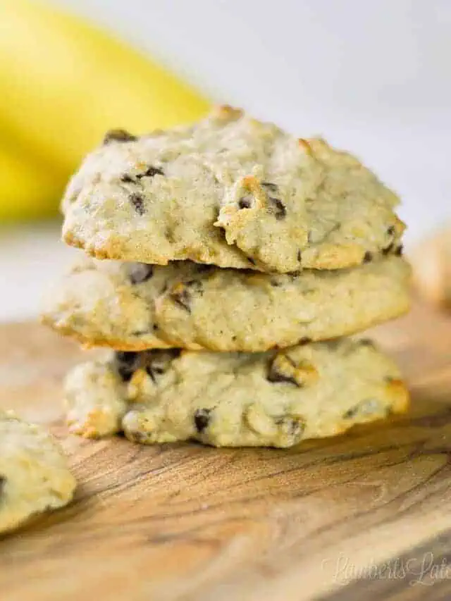 three banana bread chocolate chip cookies stacked on a cutting board.