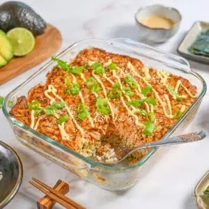 a spicy salmon sushi bake in a casserole dish, surrounded by toppings like avocado and mayo.
