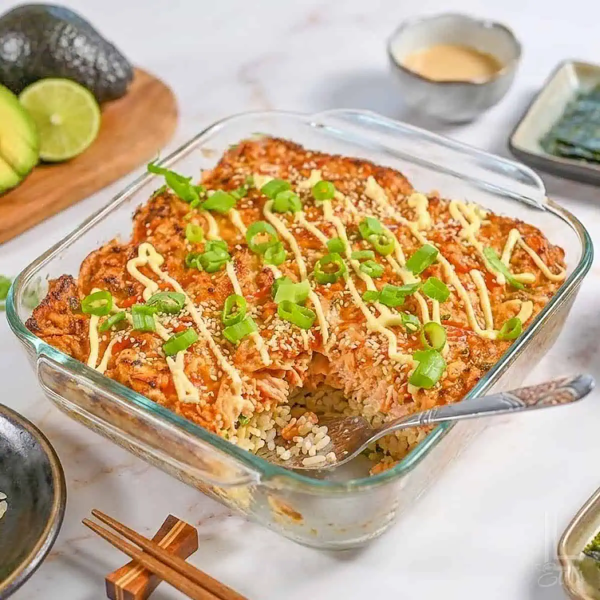 a spicy salmon sushi bake in a casserole dish, surrounded by toppings like avocado and mayo.