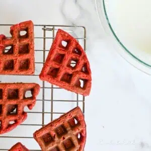 red velvet waffles on a wire cooling rack, with bowl of cream cheese glaze.