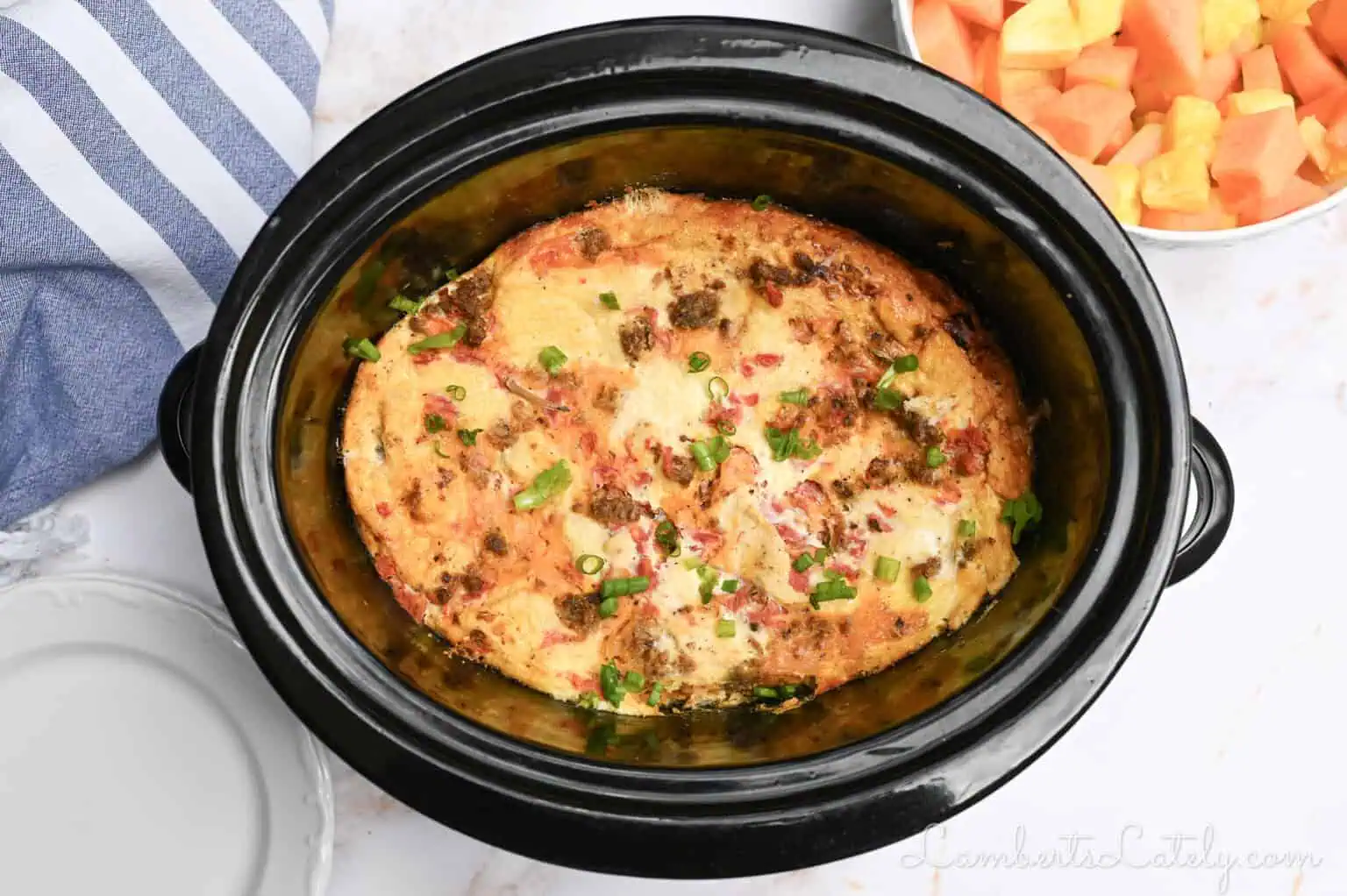 breakfast casserole in a slow cooker, with fruit, a towel, and plates surrounding it.