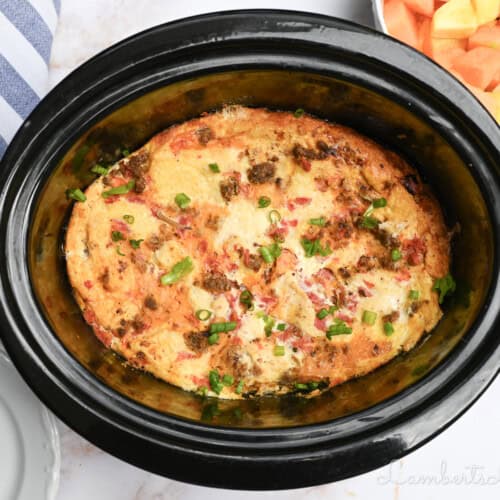 breakfast casserole in a slow cooker, with fruit, a towel, and plates surrounding it.