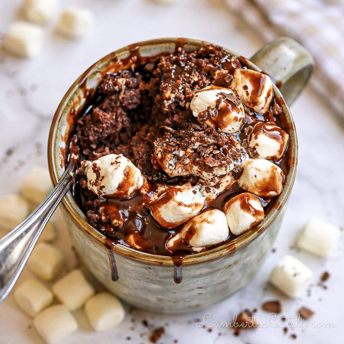hot chocolate mug cake, covered in marshmallows and chocolate syrup, with a spoon.