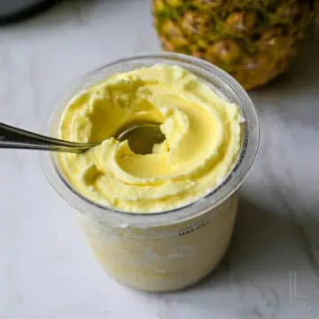 Creamy homemade pineapple dole whip soft serve in a Ninja Creami pint next to a whole fresh pineapple.