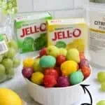 Colorful frozen sour grapes in a white bowl with lemon and Jello boxes nearby.