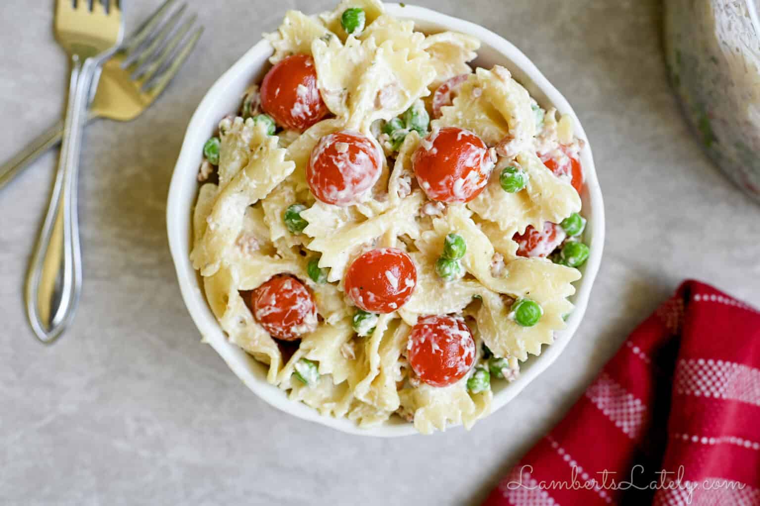 bacon ranch pasta salad in a bowl, topped with tomatoes and peas, next to forks, napkin, serving bowl.