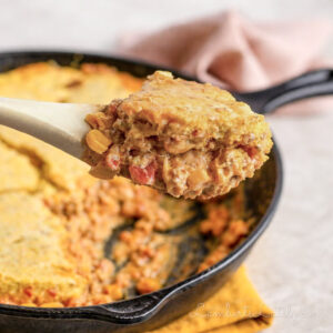 holding a spoonful of cowboy casserole topped with cornbread in front of a cast iron skillet.