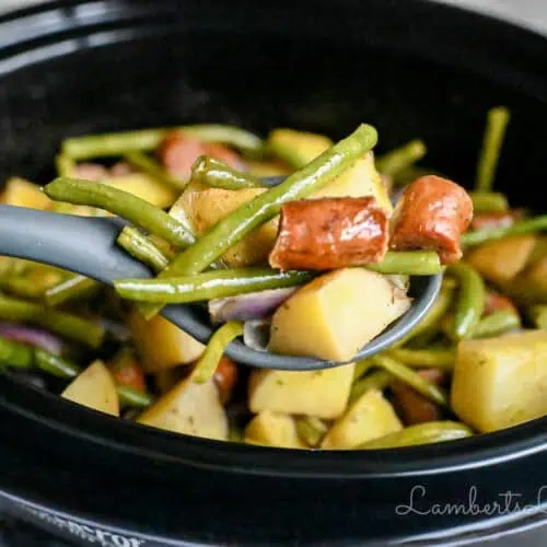 holding a spoonful of swamp potatoes with green beans and sausage in front of a crock pot.