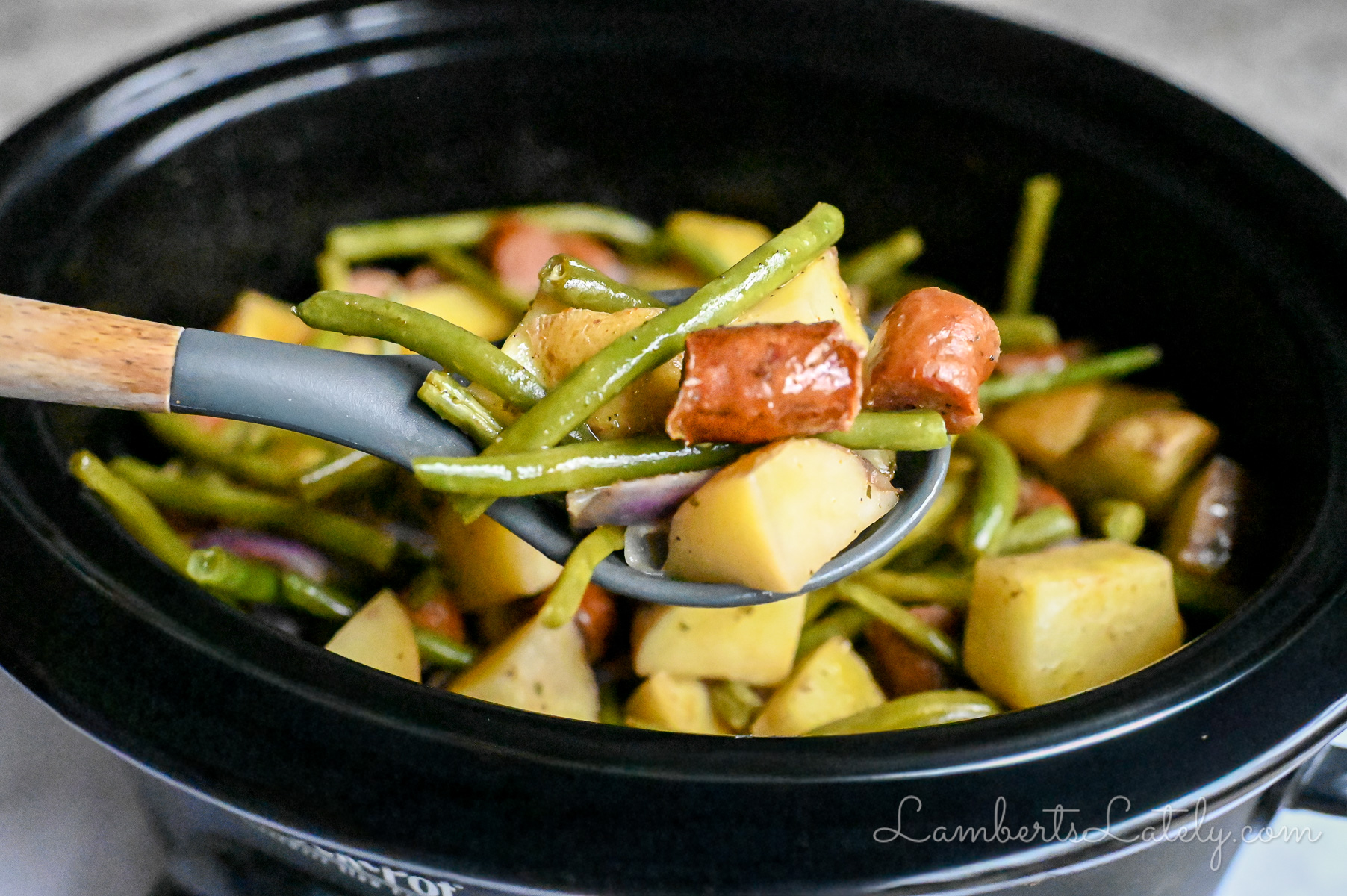 holding a spoonful of swamp potatoes with green beans and sausage in front of a crock pot.