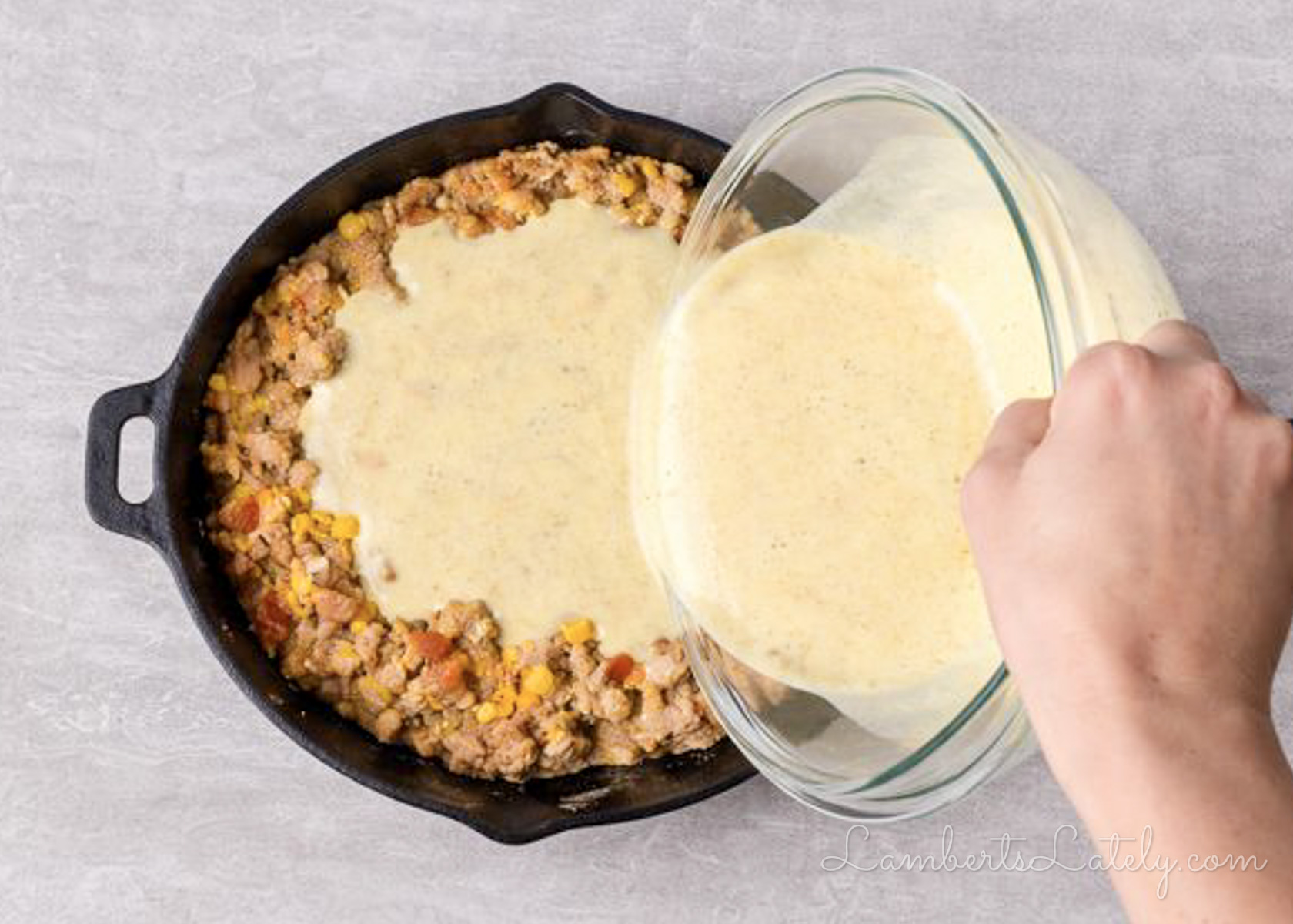 pouring cornbread batter over cowboy casserole mixture in a cast iron skillet.