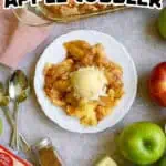 Apple cobbler served with a scoop of vanilla ice cream on a white plate surrounded by fresh apples.