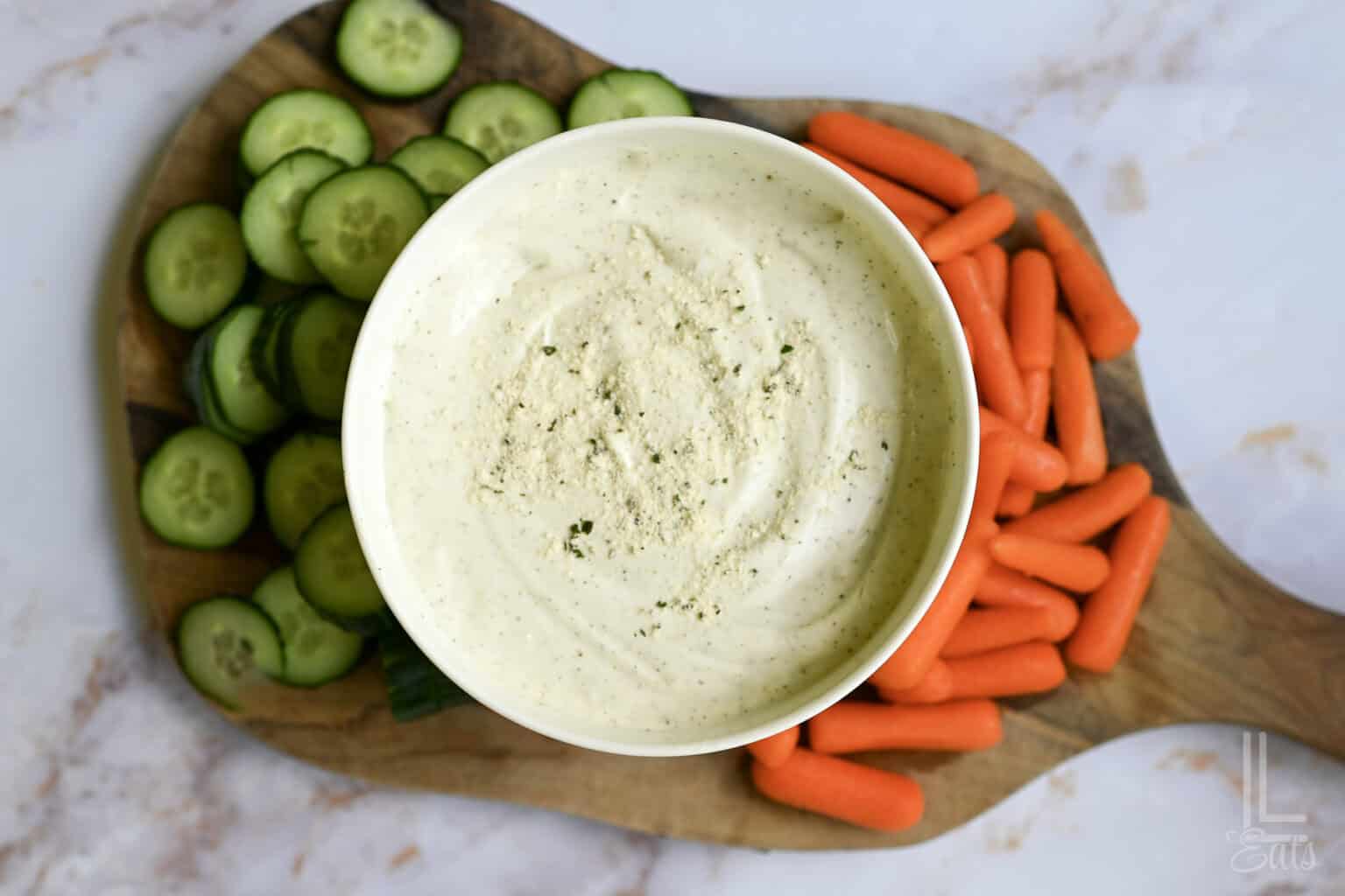 Cottage cheese ranch dip with sliced cucumbers and baby carrots on a wooden board.
