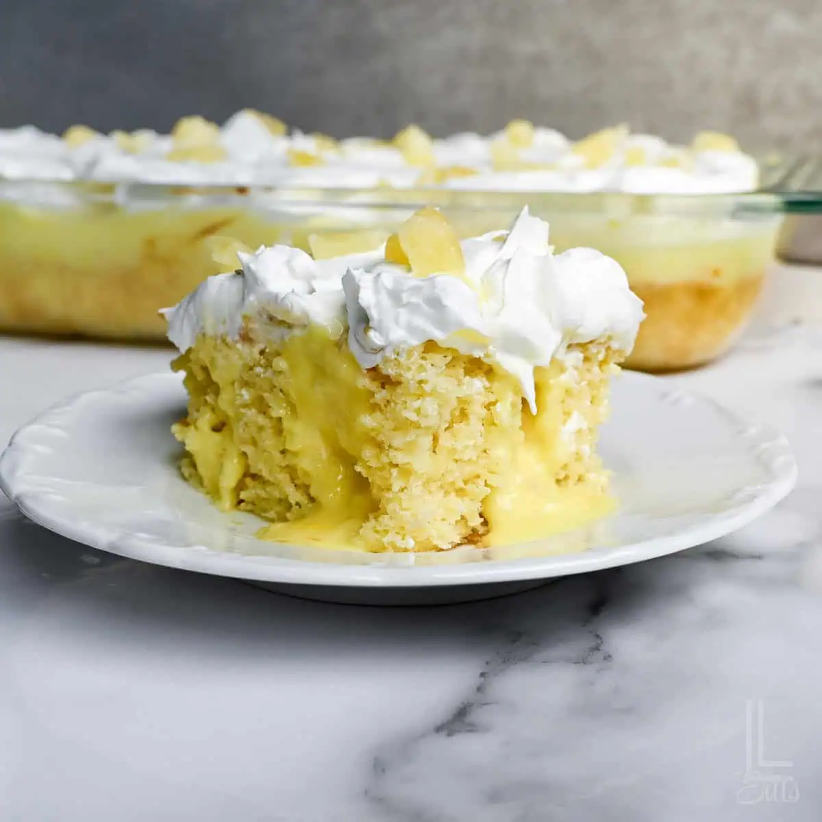 slice of pineapple poke cake, showing pineapple pudding filling, on a plate in front of a baking dish.