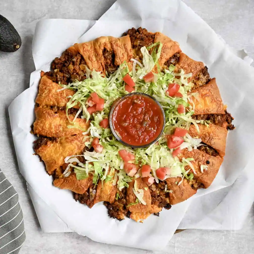 taco crescent ring with a cheesy crust on parchment paper, with lettuce and tomato, jar of salsa in the middle.