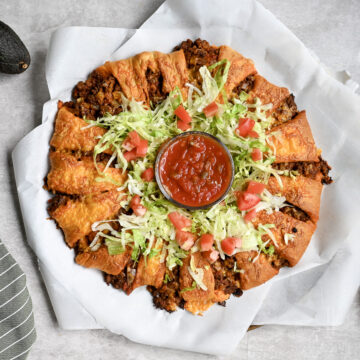 taco crescent ring with a cheesy crust on parchment paper, with lettuce and tomato, jar of salsa in the middle.