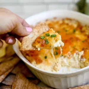 holding a bite of creamy crab rangoon dip on a wonton chip in front of a dish of dip covered in sweet chili glaze and green onions.